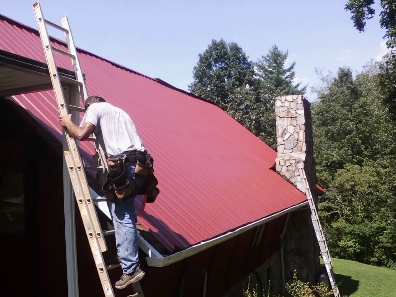 Roofer on a ladder beside a red metal roof and stone chimney — Soffit & Fascia Repair by Roofing Brothers in Knoxville